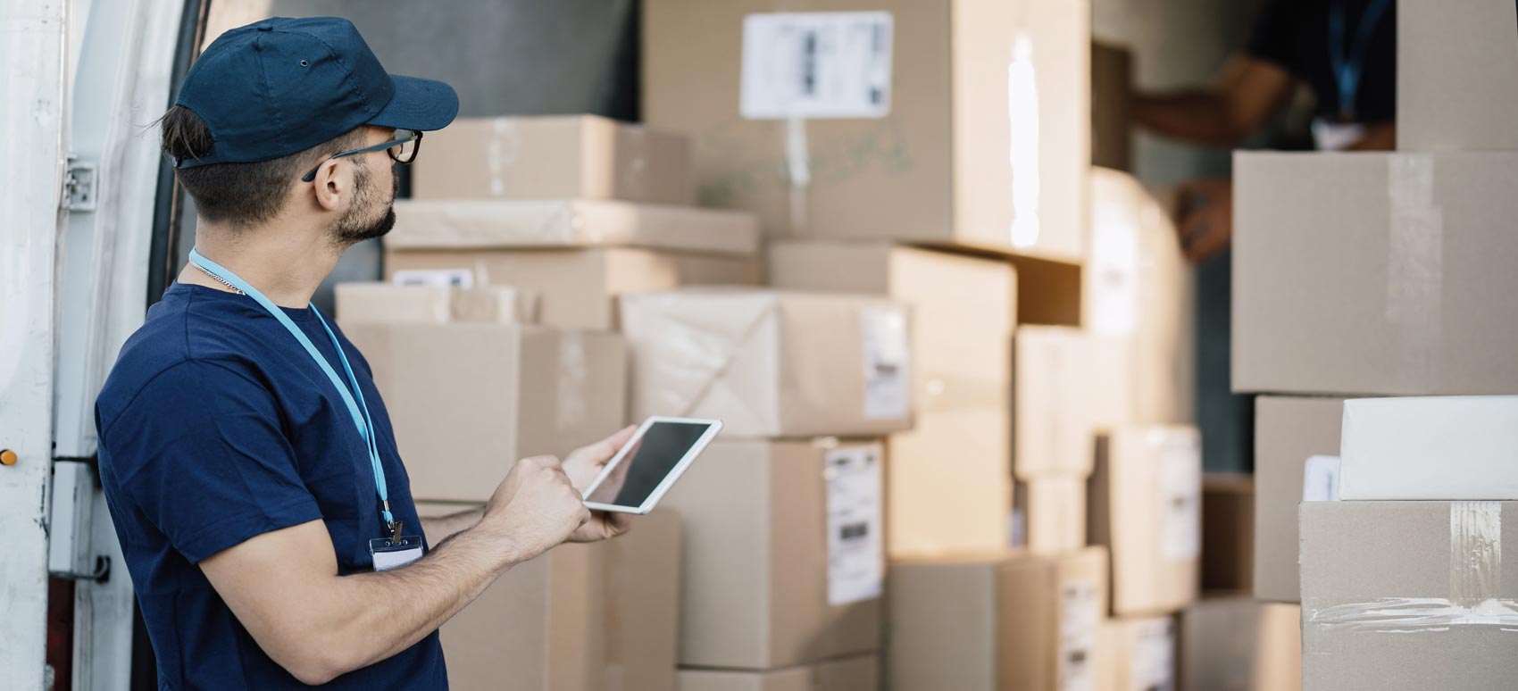A delivery worker holding a digital tablet checks and manages packages being loaded into a delivery van filled with cardboard boxes.
