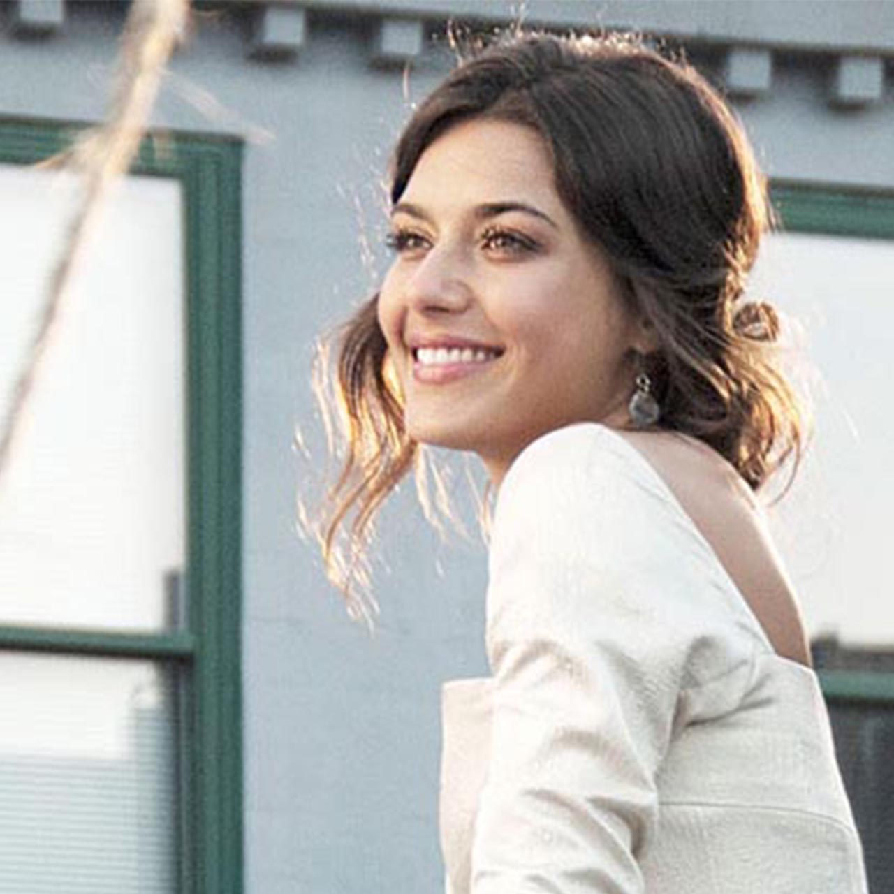 A smiling woman with styled hair, wearing a light-coloured outfit and delicate earrings, captured in soft, natural lighting