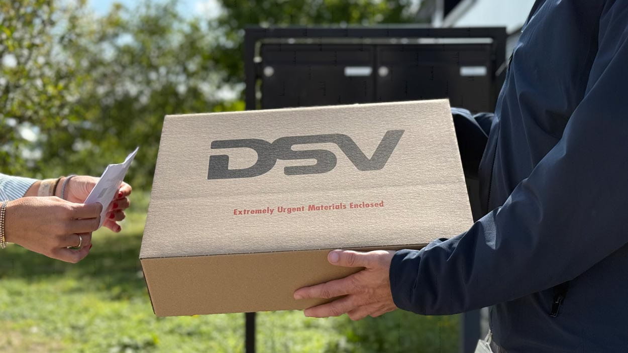 A courier hands over a DSV parcel labelled 'Extremely Urgent Materials Enclosed' to a recipient signing for delivery outdoors